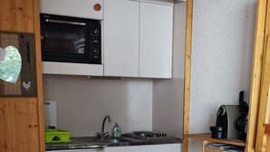 Cocina compacta con gabinetes blancos, un microondas, estufa y un lavamanos pequeño. Hay una silla de madera y un bote de basura en la esquina.