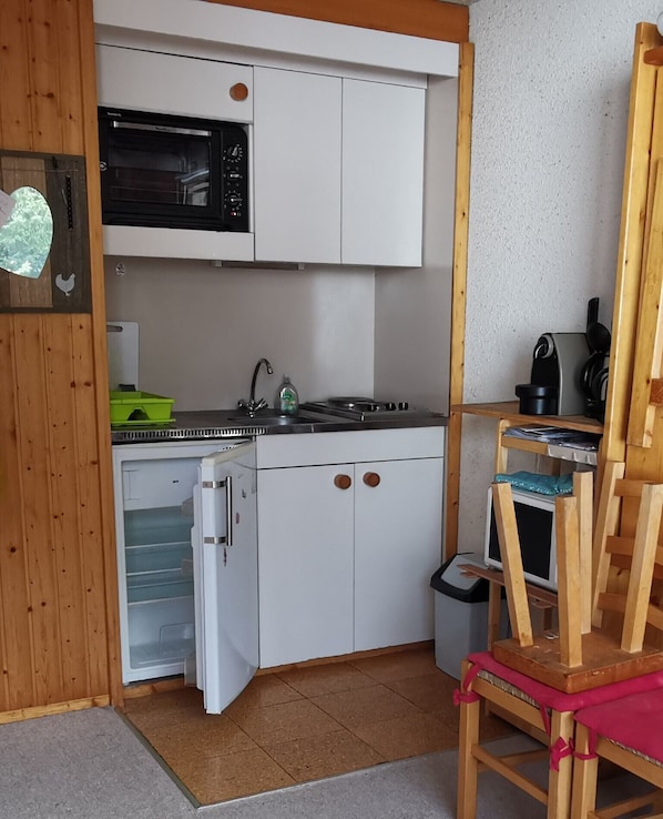 Cocina compacta con gabinetes blancos, un microondas, estufa y un lavamanos pequeño. Hay una silla de madera y un bote de basura en la esquina.