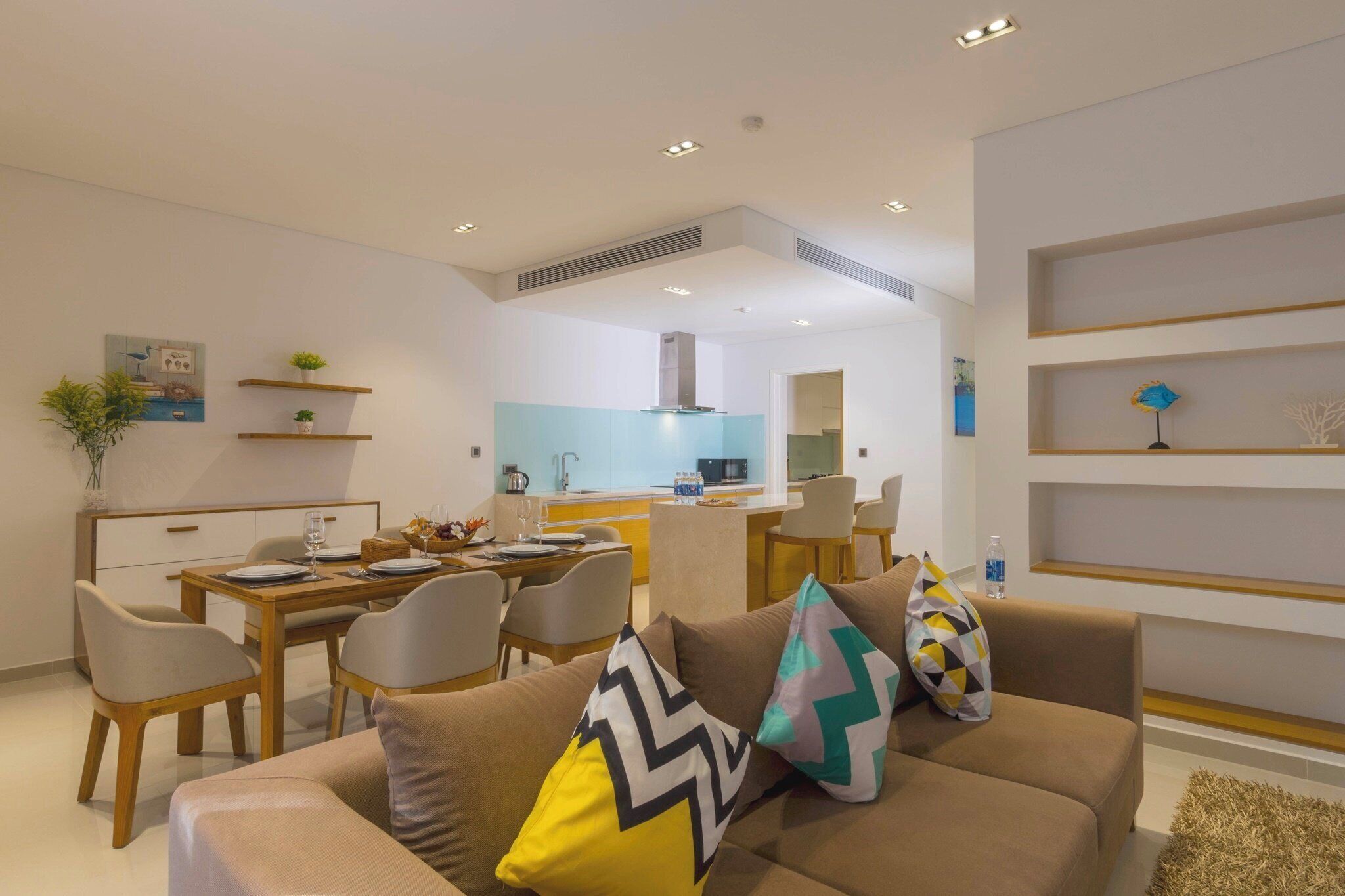 A modern living room with a sofa, dining table, and chairs. There is a kitchen area in the background with a microwave and a sink.