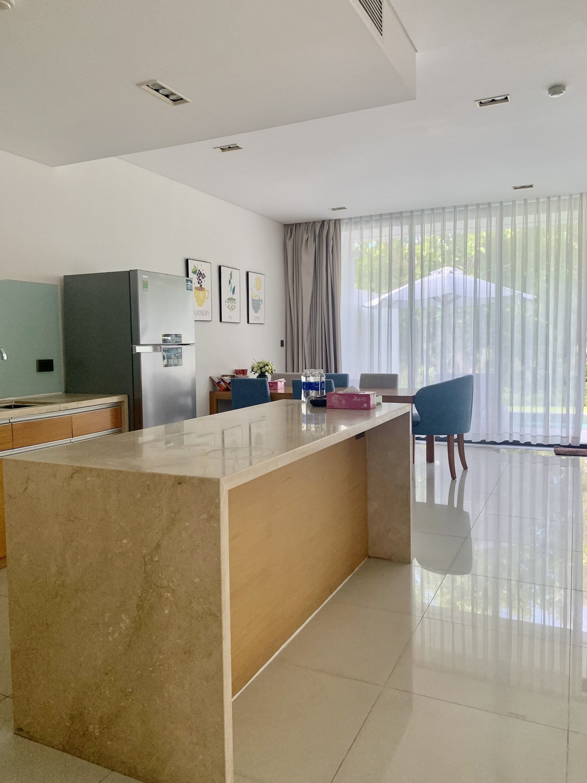 A modern kitchen with a marble island, stainless steel appliances, and a large window with sheer curtains.