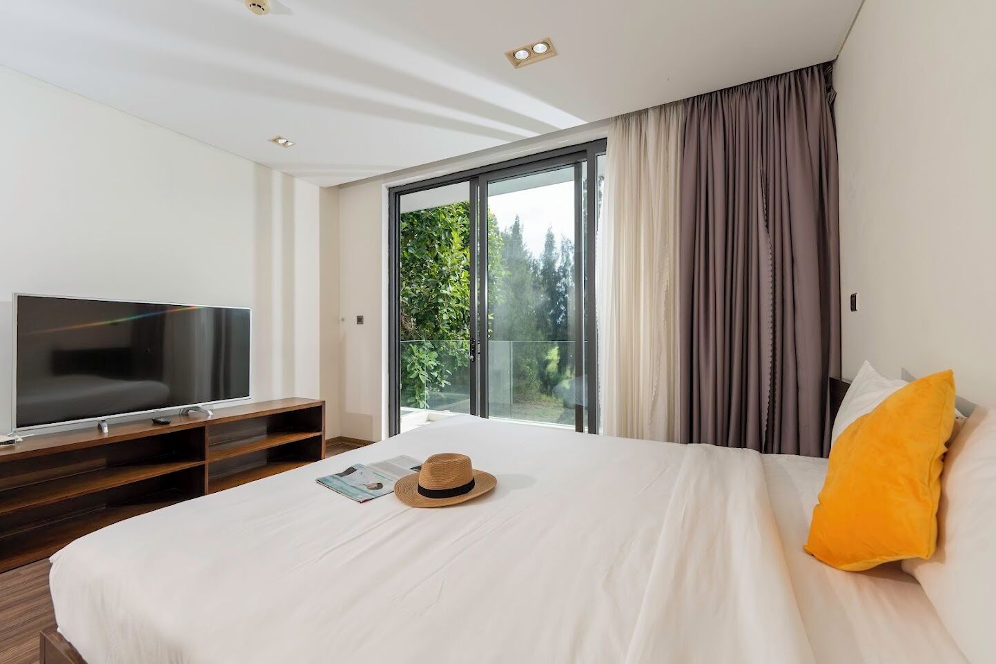 Ein modernes Hotelzimmer mit einem großen Bett, einem Flachbildfernseher, einem Balkon mit Blick auf Bäume und einer Glastür.