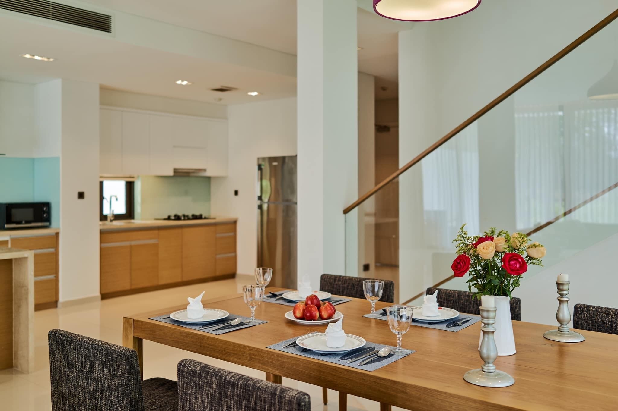 A dining area with a wooden table set for four, a vase with flowers, and a kitchen in the background.