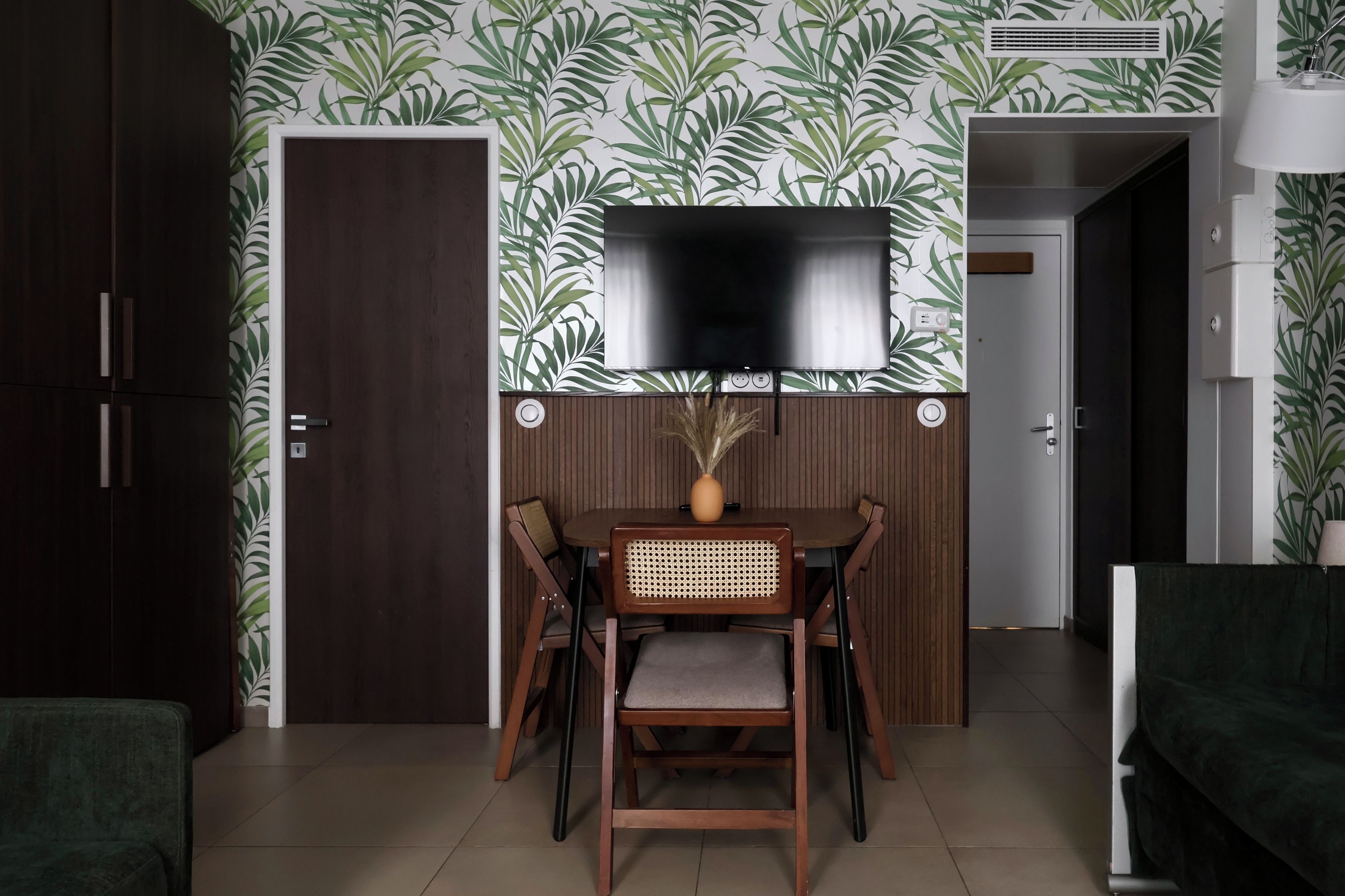 Een eetgedeelte met een aan de muur bevestigde televisie, een houten eettafel met stoelen en een deur links.