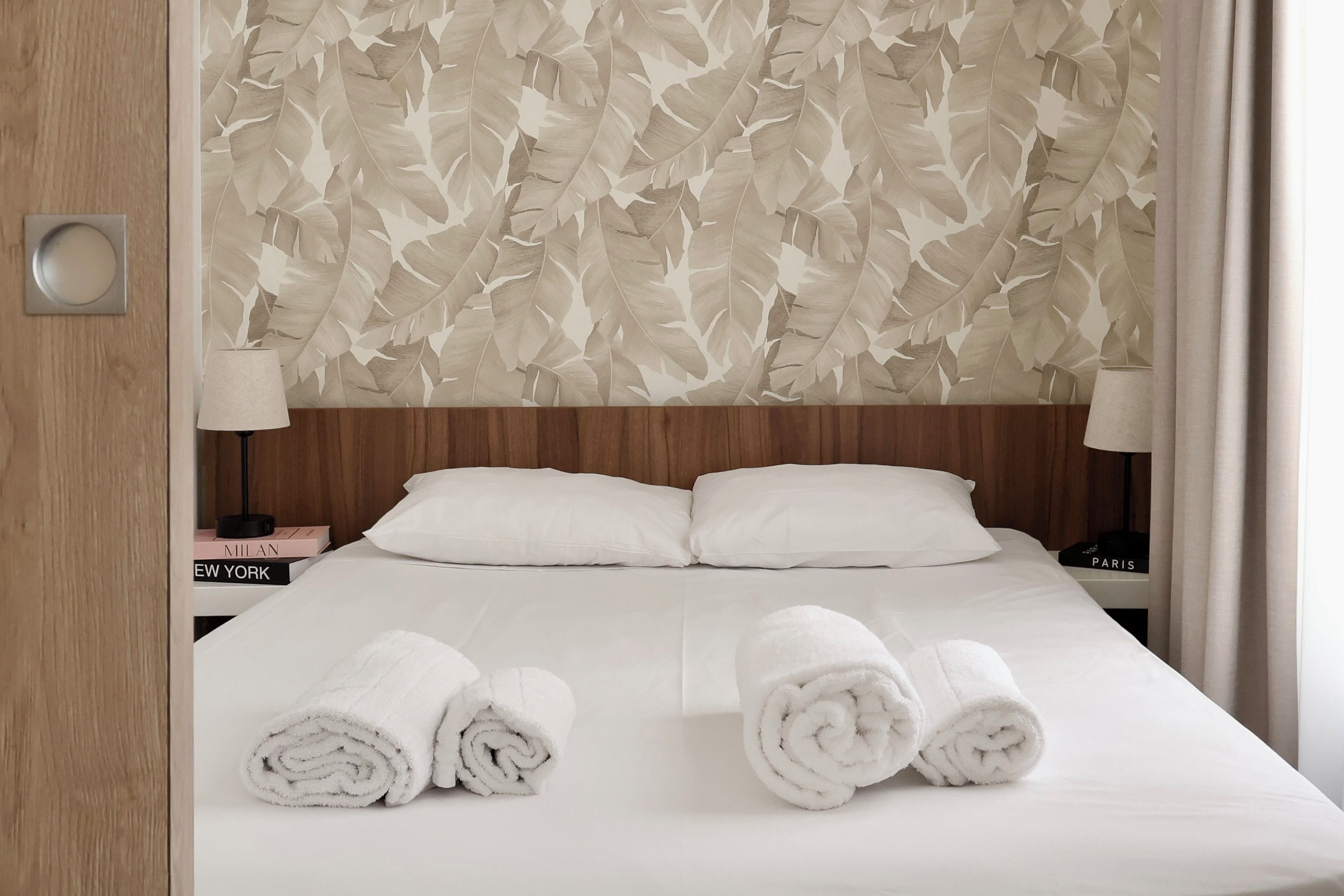 A hotel room with a bed, two pillows, two bedside tables with lamps, and two rolled towels on the bed.