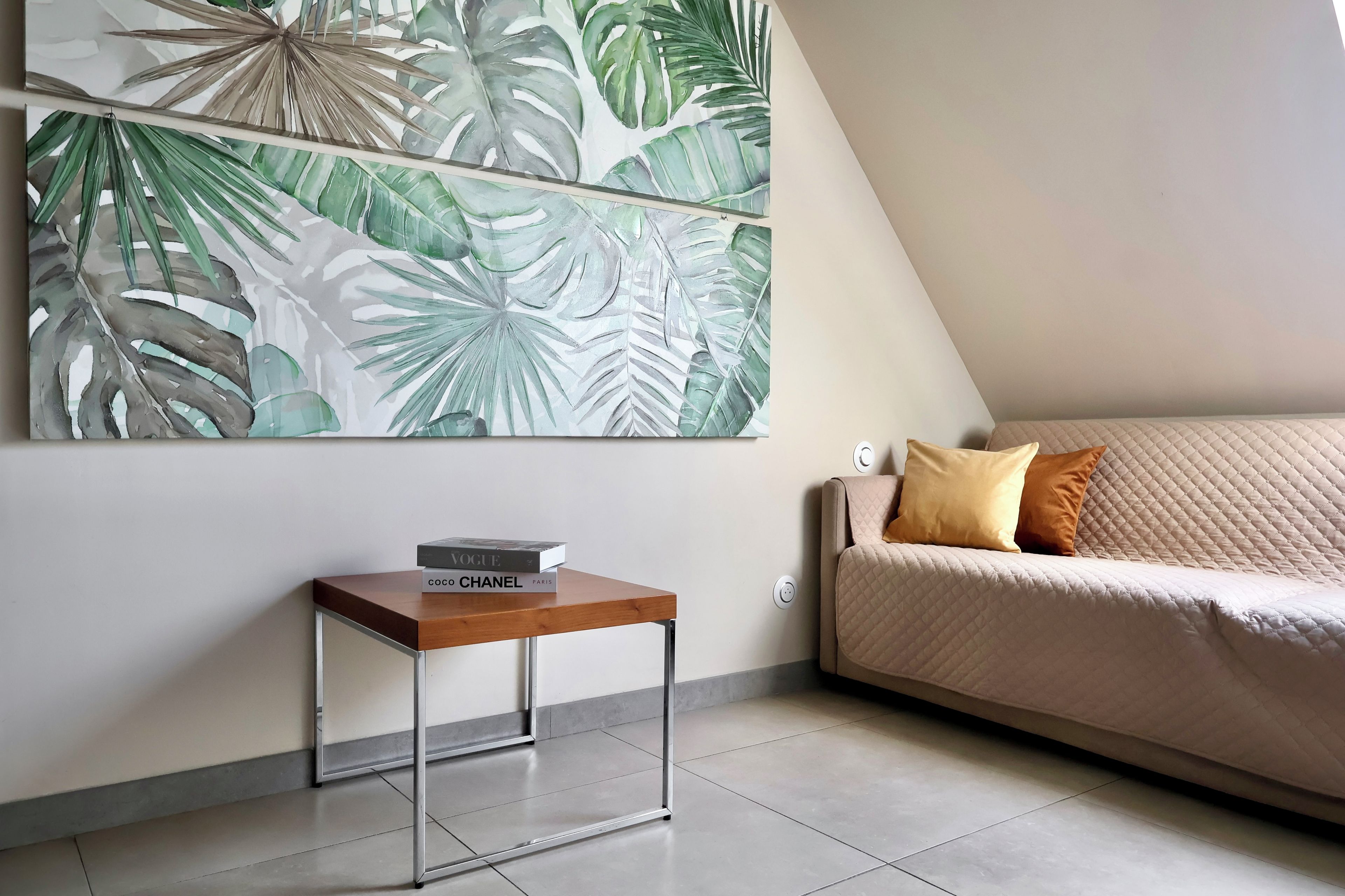 A modern living room with a large tropical leaf painting, a small wooden side table with a book, and a beige sofa with a quilted cover.