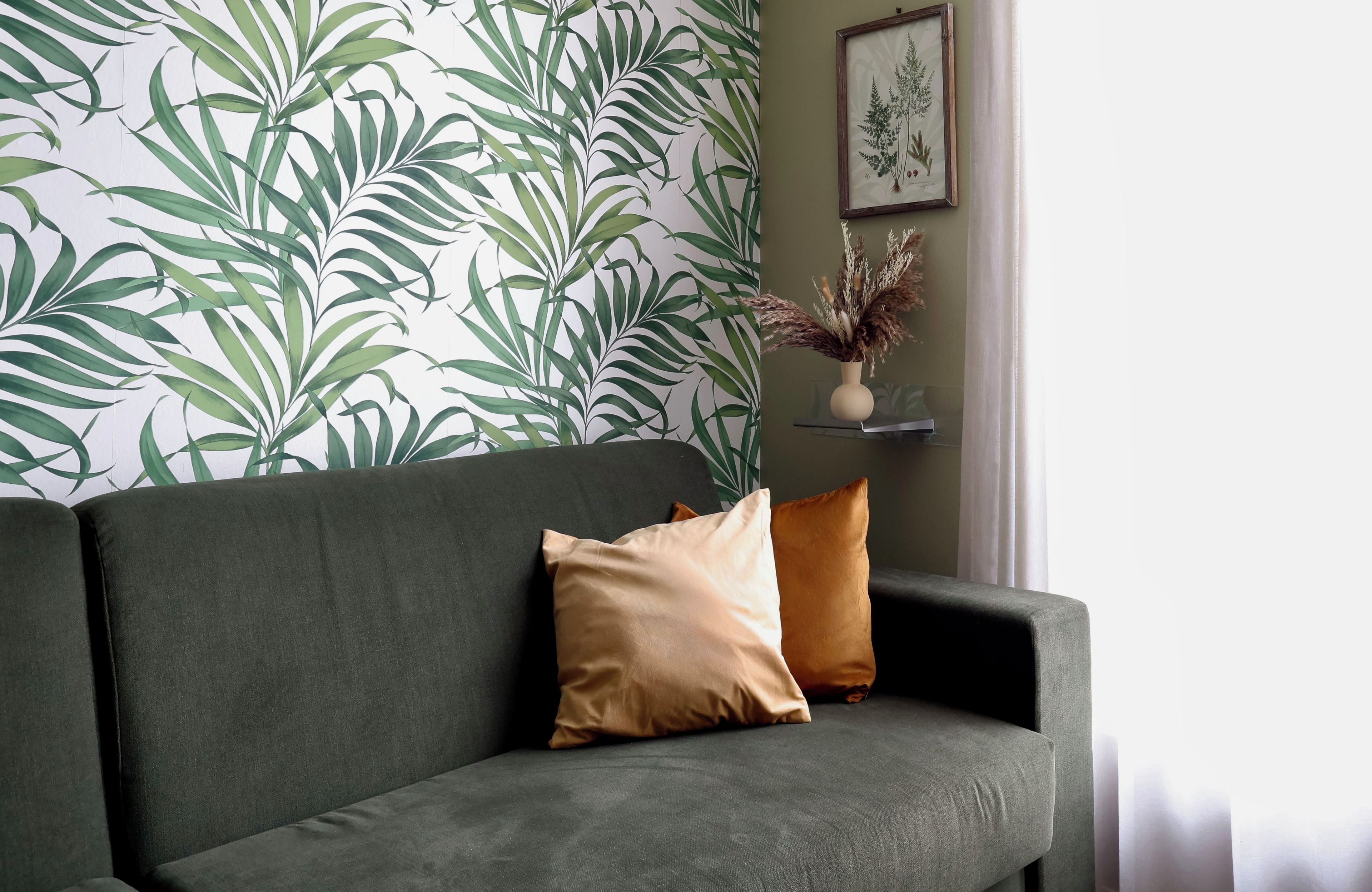 Een groene bank met twee kussens, een muur met een bladerenprint en een ingelijste botanische tekening aan de muur.