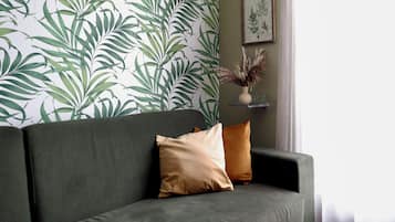 Een groene bank met twee kussens, een muur met een bladerenprint en een ingelijste botanische tekening aan de muur.