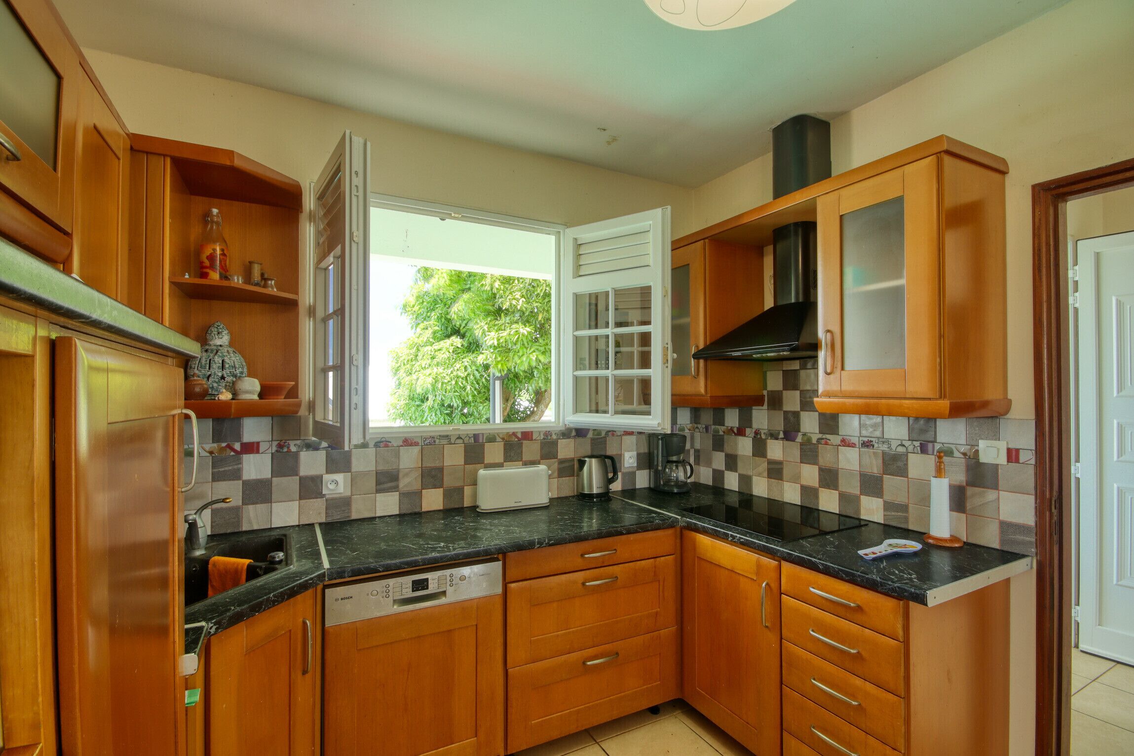 A kitchen with wooden cabinets, a black countertop, and a window with a view of greenery outside.