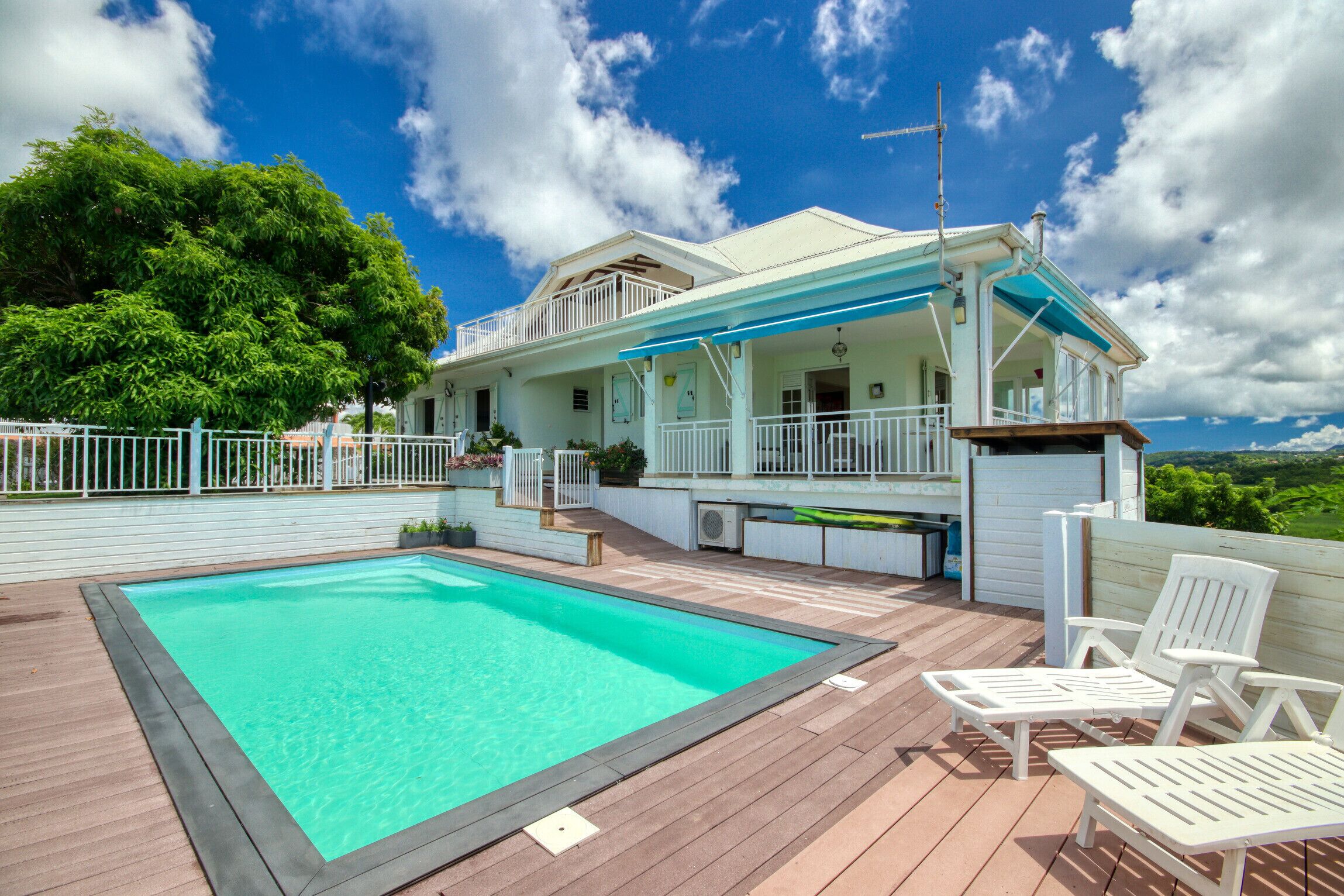 A poolside area with a clear pool, white deck chairs, and a house with a balcony overlooking the landscape.