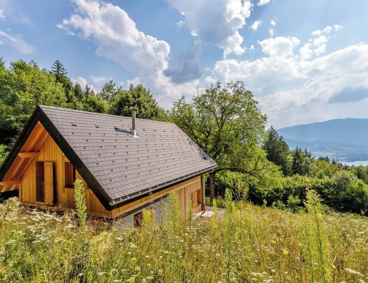 A wooden cabin with a sloped roof, surrounded by lush greenery and a mountainous backdrop.