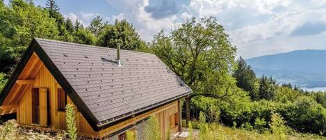 Une cabane en bois, dotée d’un toit en pente, entourée d’une végétation luxuriante et avec une chaîne de montagnes en arrière-plan.