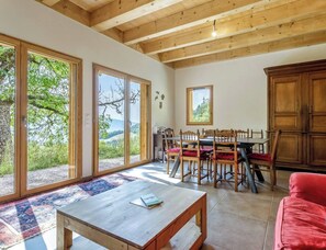 Amplio salón con una mesa de comedor de madera, sillas y un sofá rojo. Grandes ventanales ofrecen vistas a zonas verdes y colinas.