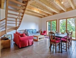 Amplia sala de estar con escalera de madera, un sofá gris, un sofá rojo, una mesa de comedor con sillas y grandes ventanales con vistas a zonas verdes.
