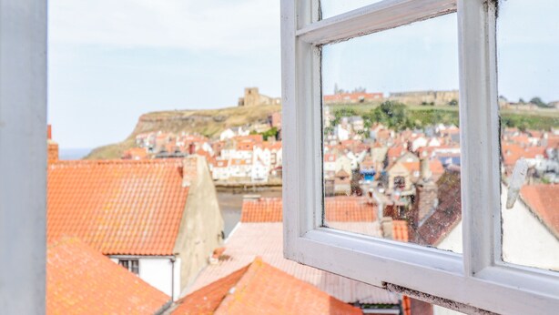 En vy genom ett fönster som visar en stad med röda tak och en kulle med en byggnad på toppen.