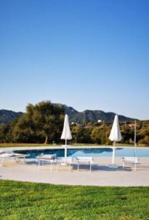 Piscina com espreguiçadeiras e guarda-sóis, cercada por gramado e montanhas ao fundo.