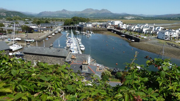 A marina with boats docked, a bridge, and buildings in the background. - Wenallt (Porthmadog)