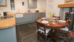 A modern kitchen with grey cabinets, a wooden dining table set for four, and a wood-burning stove. - Wenallt (Porthmadog)