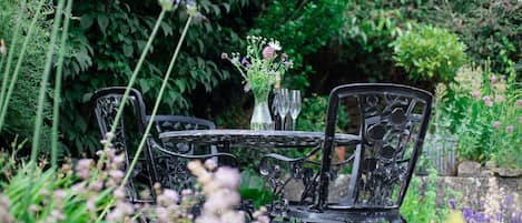 A garden setting with a round table and four chairs, surrounded by lush greenery and flowering plants.