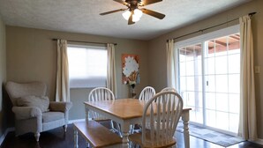 Une salle à manger avec une table en bois et des chaises blanches, un ventilateur de plafond et de grandes fenêtres avec des rideaux.
