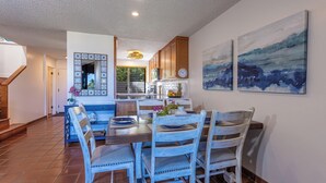 A dining area with a wooden table and chairs, a kitchen in the background, and two abstract paintings on the wall. - Townhouse Retreat with Ocean Views From Upper & Lower Lanai - Wailea Ekahi #30B (Wailea, Maui)