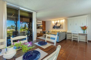 Ein Essbereich mit Holztisch, Stühlen und Blick ins Freie. Im Wohnbereich befindet sich eine Couch und ein kleiner Tisch mit einem Blumenstrauß.