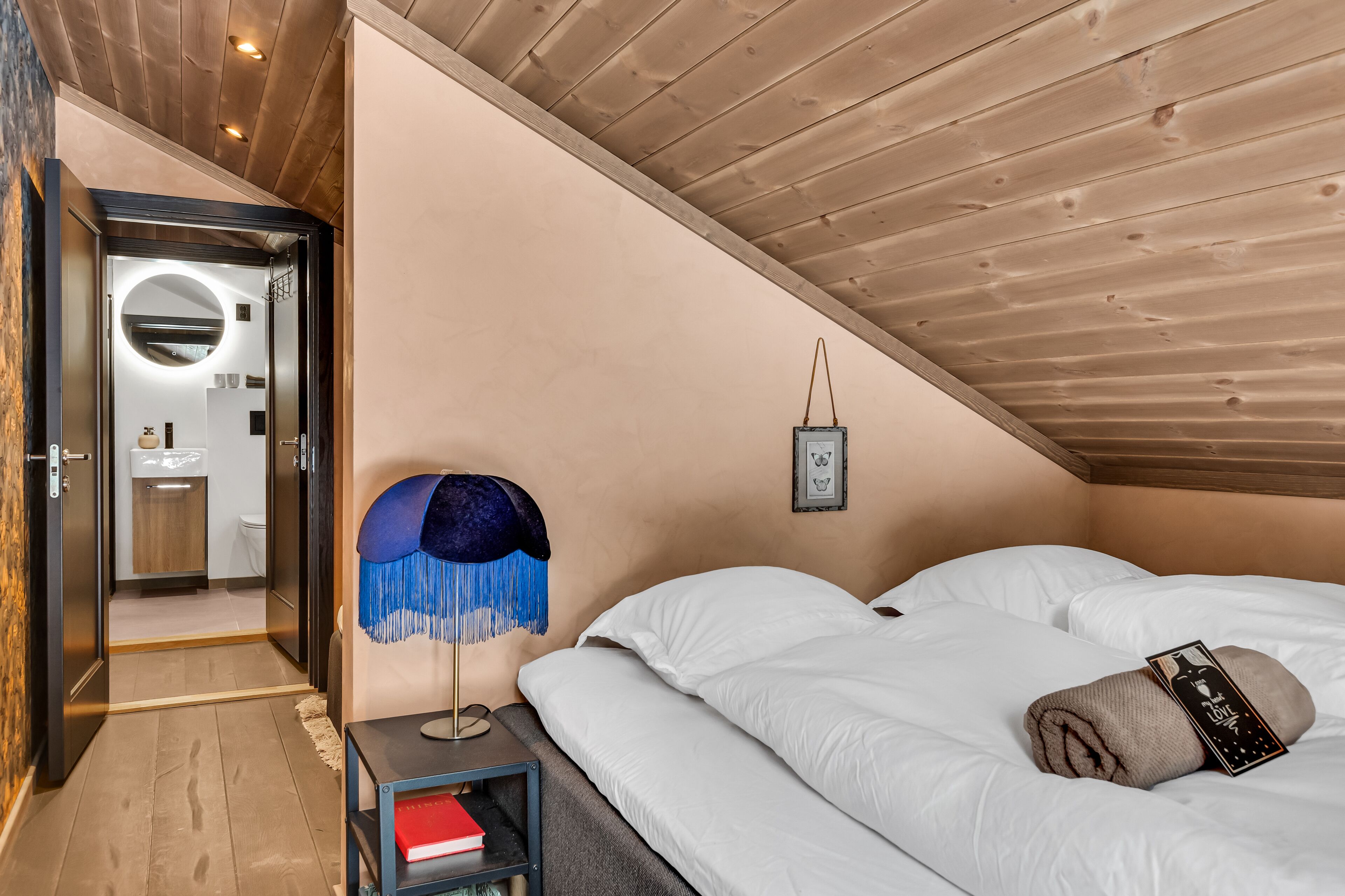 A bedroom with a wooden ceiling, a bedside table with a lamp, and a bed with white bedding.