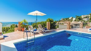 A swimming pool with lounge chairs and an umbrella, surrounded by a scenic view of the sea and hills. - Villa Estrella with private heated pool and breathtaking views (AN)