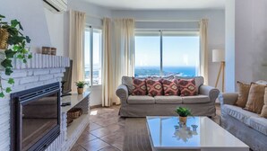 A living room with a fireplace, a sofa with patterned cushions, a coffee table, and a view of the ocean. - Villa Estrella with private heated pool and breathtaking views (AN)