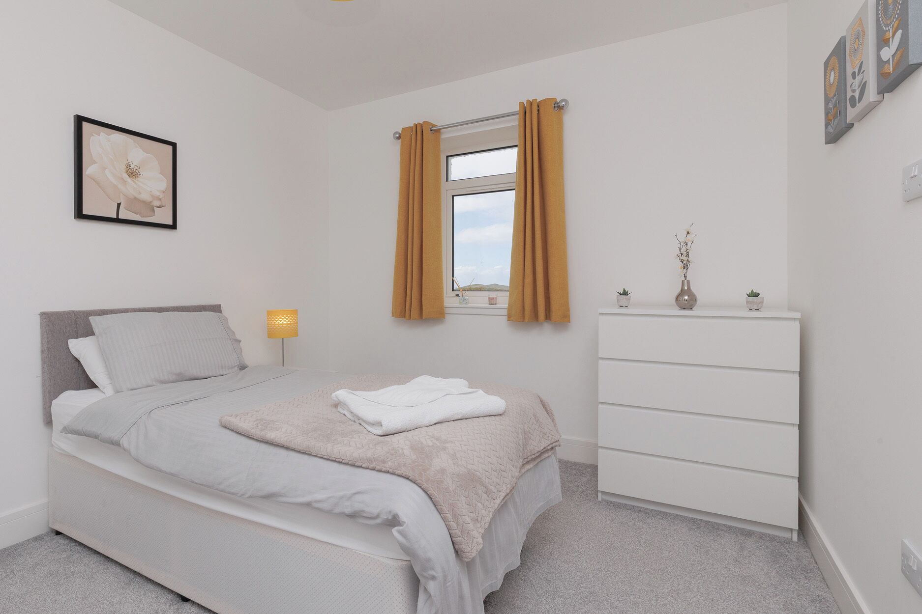 A bedroom with a bed, a dresser, a window with curtains, a bedside lamp, and a framed picture on the wall.