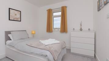 A bedroom with a bed, a dresser, a window with curtains, a bedside lamp, and a framed picture on the wall.