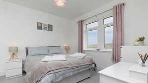 A bedroom with a bed, bedside tables, a desk, and a window with curtains. - Dun Buidhe (South Uist)