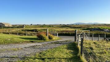 A rural landscape with a gravel path leading to a gated entrance, surrounded by grassy fields and scattered farm buildings.
