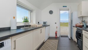 A compact kitchen with white cabinets, a black countertop, a microwave, and a dishwasher. There is a fire exit sign above the door, and a fire extinguisher mounted on the wall. - Dun Buidhe (South Uist)