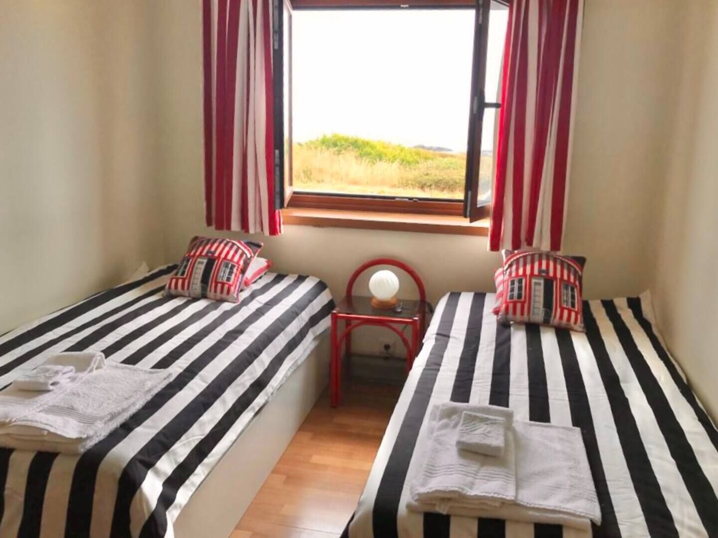 Una habitación con dos camas, cada una con ropa de cama a rayas y almohadas a juego. Hay una mesita con una lámpara y una ventana con cortinas.