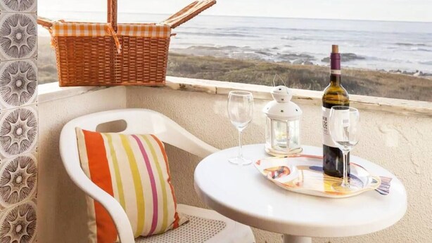 A balcony with a wicker basket, a white chair with a striped cushion, a small table with a wine bottle, glasses, and a lantern. - Beach's House em Moledo (Moledo)