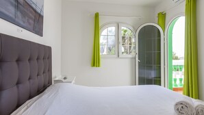 A bedroom with a tufted headboard, a bed with white linens, a bedside table with a lamp, and a window with green curtains.
