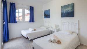 A hotel room with two single beds, a window with curtains, a bedside table with a lamp, and artwork on the wall.