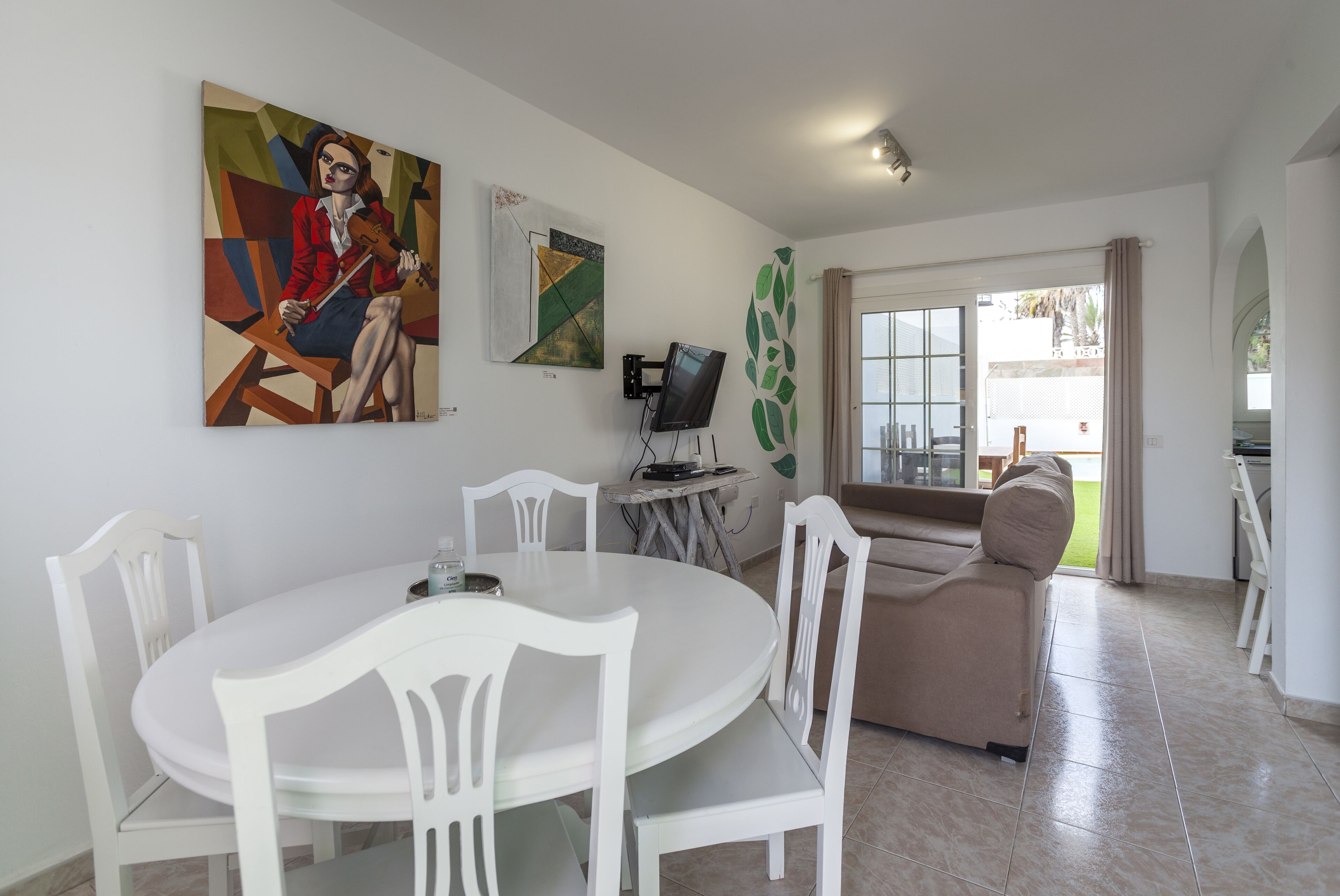 Zona pranzo con tavolo rotondo e sedie bianche, un divano, una televisione e opere d'arte alle pareti.