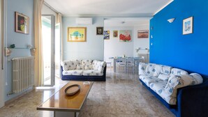 A living room with a blue wall, a white sofa, a wooden coffee table, and a dining area with a table and chairs. - 2419 Appartamento Kalepolis (Gallipoli)