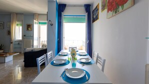 A dining area with a white table set for four, blue placemats, and a view of the balcony through the window. - 2419 Appartamento Kalepolis (Gallipoli)