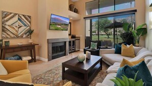 A spacious living room with a fireplace, comfortable seating, and large windows offering a view of the outdoors. - Fabulously Remodeled 2 Bedroom Condo With Beautiful Golf and Mountain Views (La Quinta)