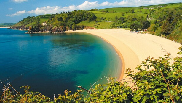A coastal landscape with a sandy beach, clear blue waters, and green hills in the background. - Crew House (Dartmouth)