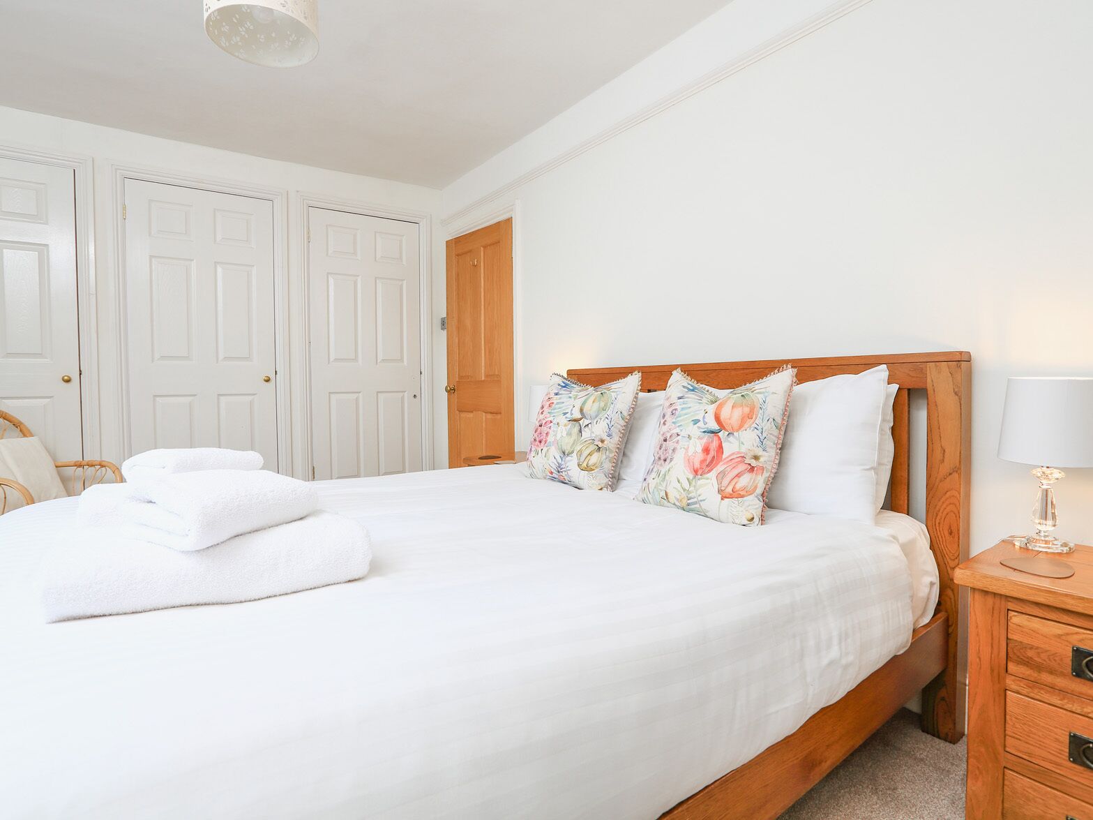 A neatly made bed with white linens and floral pillows, a wooden nightstand with a lamp, and a wicker chair.