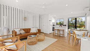 A modern, open-plan living space with a kitchen, dining area, and large windows offering a view of the outdoors. - Bella Vita by Experience Jervis Bay (Vincentia)