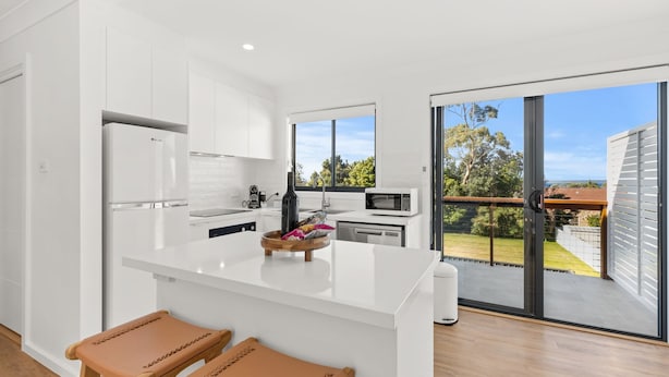 A modern kitchen with white cabinetry, a central island with a wooden bowl, and a view of a garden through large sliding glass doors. - Bella Vita by Experience Jervis Bay (Vincentia)