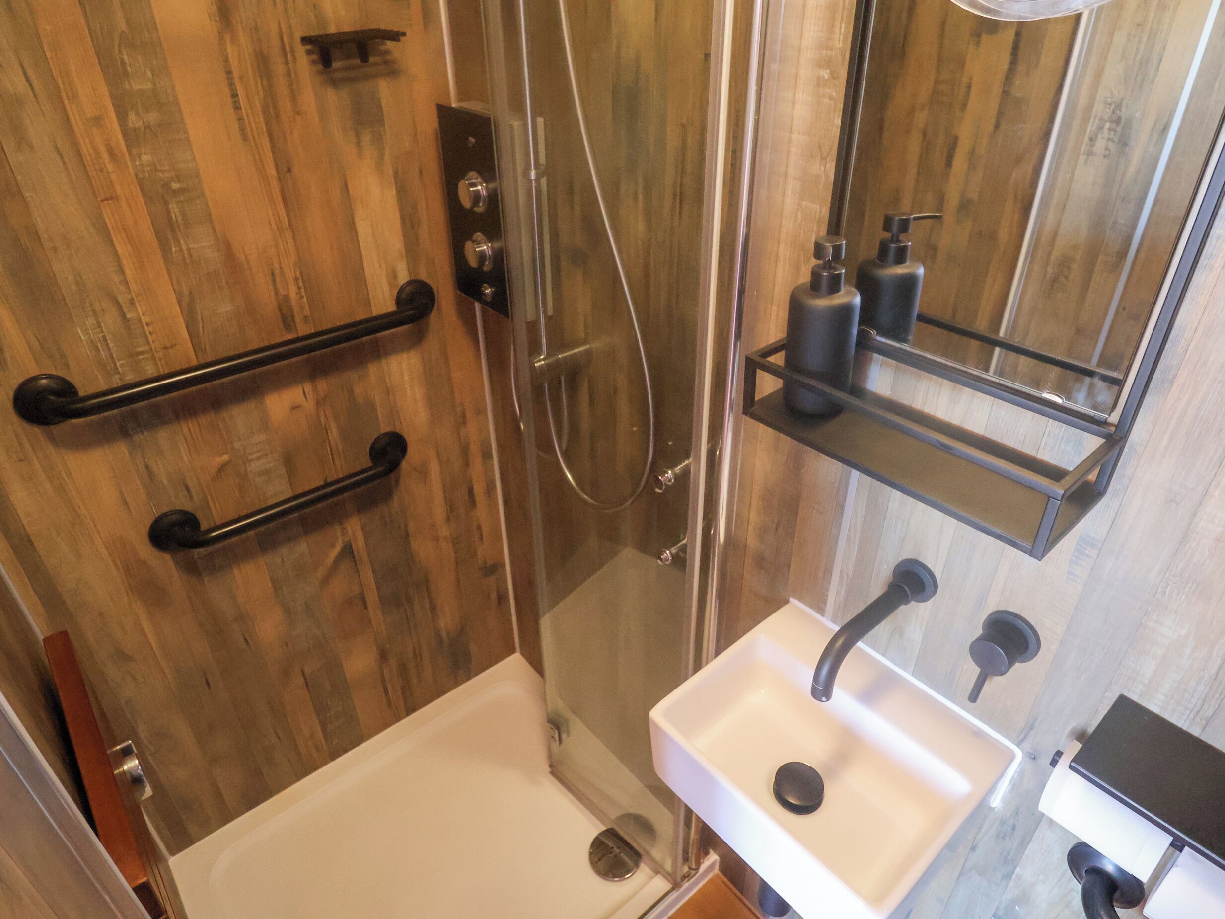 Un baño con pared de madera, cabina de ducha de vidrio, lavamanos blanco y espejo sobre el lavamanos.