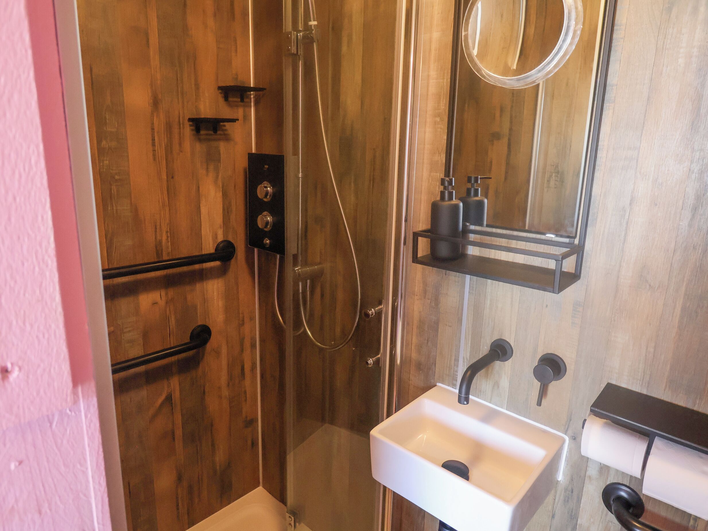 A bathroom with a wooden wall, a glass-enclosed shower, a white sink, and a mirror.