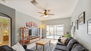 Ein Wohnzimmer mit einer dunkelgrauen Sofagarnitur, einem Holztisch, einem Deckenventilator und einem Fernseher auf einem Holzständer.