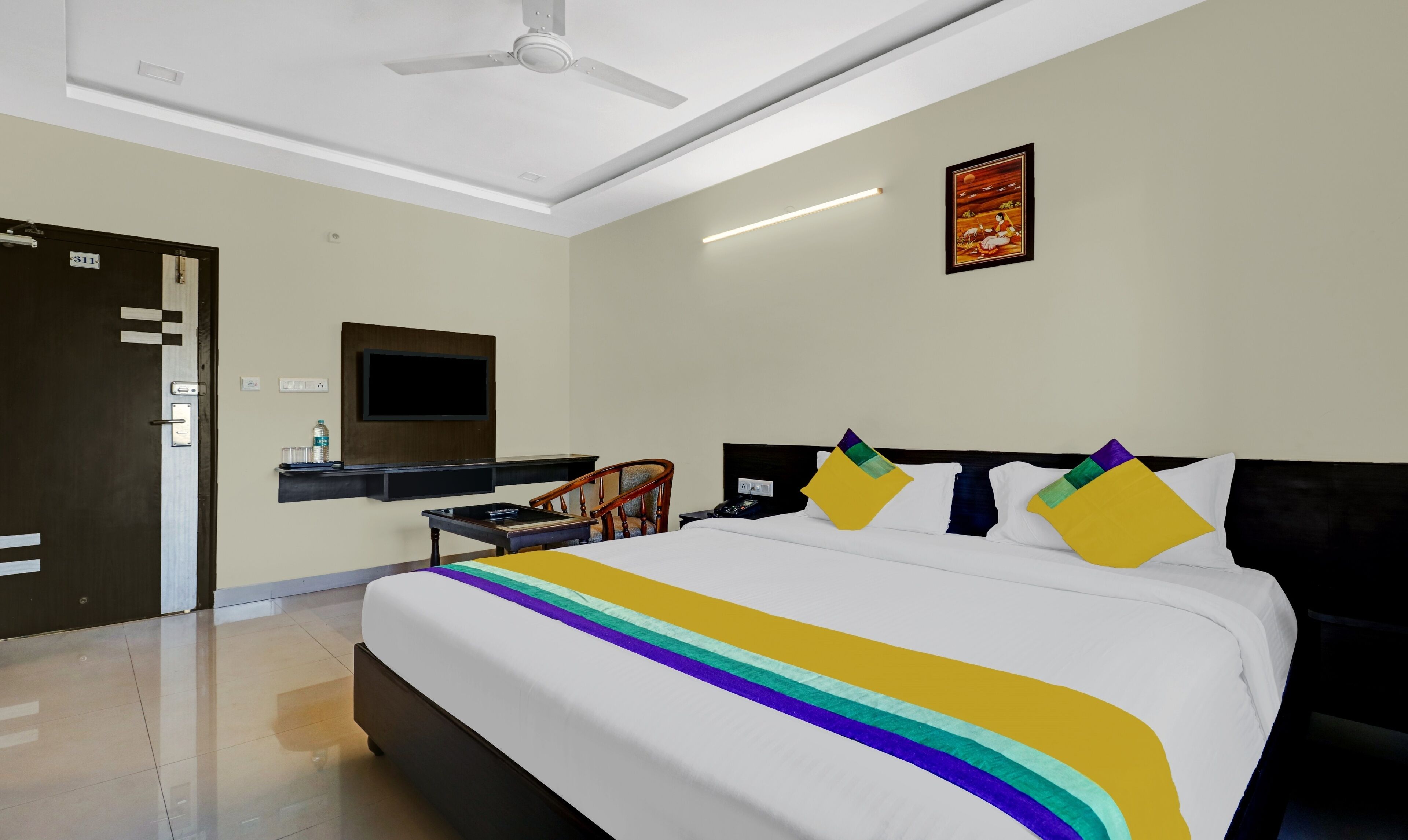 Habitación de hotel con cama, escritorio, silla, televisión montada en la pared y ventilador de techo.