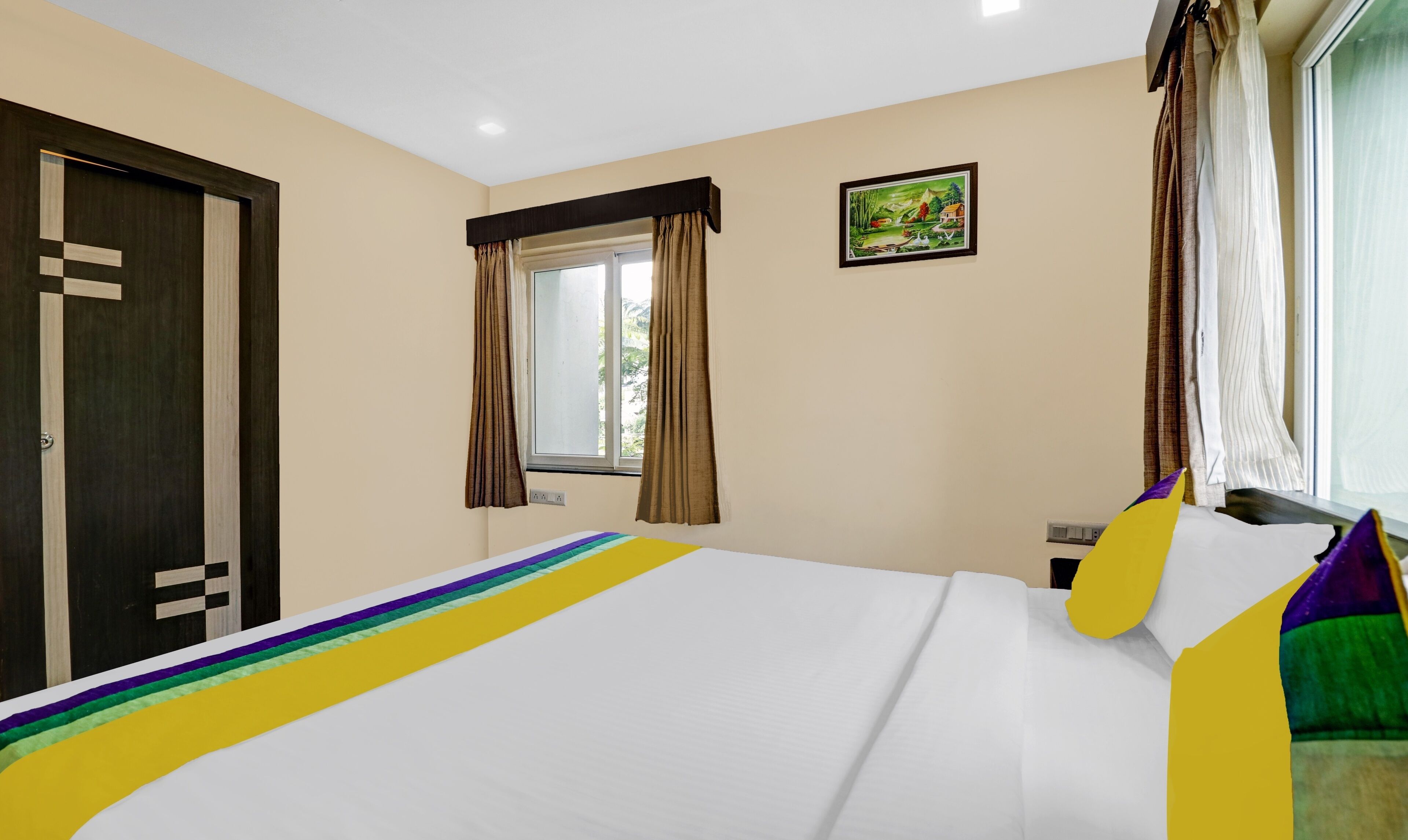 A hotel room with a bed, a window with curtains, a painting on the wall, and a door with a geometric design.